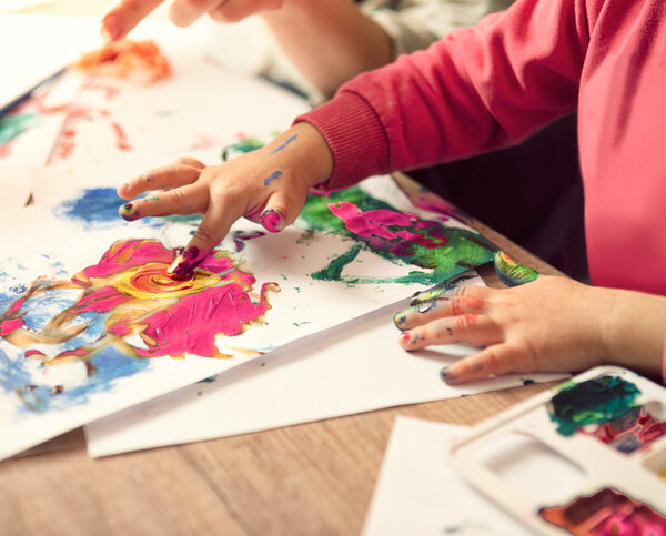 A small child in a pink shirt is shown with paint on their fingers, adding paint to paper.