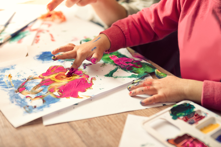 A small child in a pink shirt is shown with paint on their fingers, adding paint to paper.